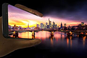 A hand holds a mobile phone in the picture, on it, the old bridge over the Main with skyline, skyscrapers in the banking district in the evening, sunset, Frankfurt am Main, Hesse, Germany, Europe