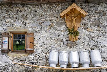 Milk cans lined up on a wooden bench, window and shrine with Jesus on the cross, facade made of rough stones from an alpine pasture, Gramaialm, Alpenpark Karwendel, Tyrol, Austria, Europe