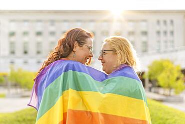 Lesbian women couple in love embracing each other, wrapped in a gay flag, in a park at sunset, with reflections of the sun. Concept of diversity, pride, love, equality