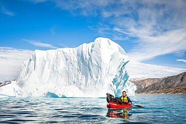 Man with packraft on fjord, icebergs and mountains behind, blue sky, Greenland, North America