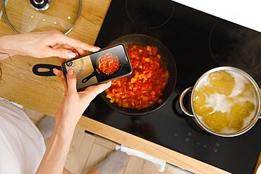 View from above of unrecognizable woman with smartphone taking photo of frying pan with tomato sauce for culinary blog while cooking in kitchen at home. Food blogger recording video of cooking process