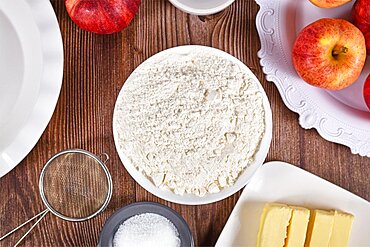 Bowl of flour surrounded by other pie crust ingredients like butter and sugar on wooden background