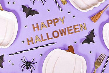 Happy Halloween text surrounded by pumpkin shaped plates, witch brooms, spiders and bats on purple background