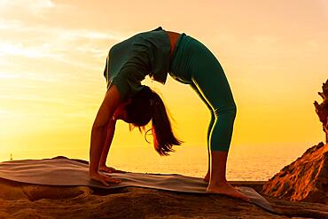 Urdhva Dhanurasana, detail of a woman doing meditation and yoga exercises on a rock at sunset next to a lighthouse in the sea, healthy and naturist life