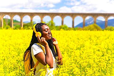 Listening to music in yellow headphones with closed eyes, a black ethnic girl with braids, a traveler, in a field of yellow flowers