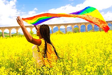 A black ethnic girl with braids holding the LGBT flag in a field of yellow flowers