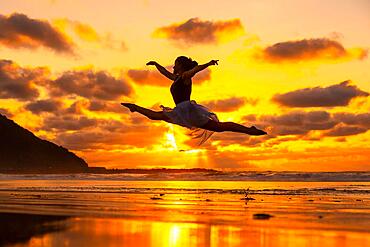 Young dancer on the beach at sunset performing a jump with the sea in the background, with the sun reflected in the water