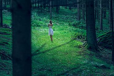 Beautiful young woman wearing elegant white dress standing in the forest with rays of sunlight beaming through the leaves of the trees