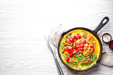 Healthy frittata in cast iron pan with fried beaten eggs and seasonal vegetables on white rustic wooden background. Italian omelette with organic spinach, bell pepper, tomatoes, top view, copy space