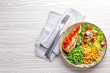 Baked chicken breast on plate with fresh salad, green peas and corn, white wooden background, top view. Healthy fitness meal with chicken fillet, balanced in proteins and carbs. Overhead, copy space