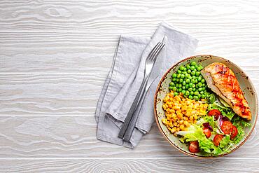 Baked chicken breast on plate with fresh salad, green peas and corn, white wooden background, top view. Healthy fitness meal with chicken fillet, balanced in proteins and carbs. Overhead, copy space