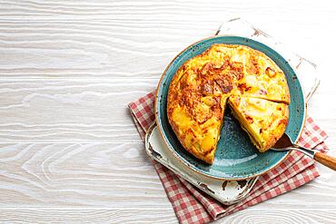 Homemade Spanish tortilla with one slice cut - omelette with potatoes on plate on white wooden rustic background top view. Traditional dish of Spain Tortilla de patatas for lunch or snack, copy space