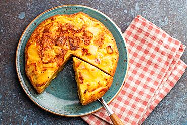 Homemade Spanish tortilla with one slice cut - omelette with potatoes on plate on stone rustic background top view. Traditional dish of Spain Tortilla de patatas for lunch or snack, overhead