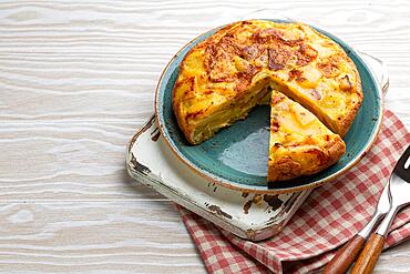 Homemade Spanish tortilla with one slice cut - omelette with potatoes on plate on white wooden rustic background top view. Traditional dish of Spain Tortilla de patatas for lunch or snack, overhead