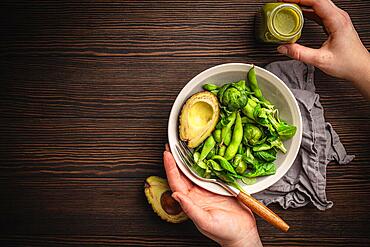 Bowl of healthy salad in female hands from above and green detox smoothie in a bottle on wooden background, top view. Woman eating tasty vegan meal, raw food diet and clean eating concept, copy space