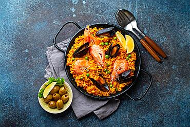 Classic dish of Spain, seafood paella in traditional pan on rustic blue concrete background top view. Spanish paella with shrimps, clamps, mussels, green peas and fresh lemon wedges from above