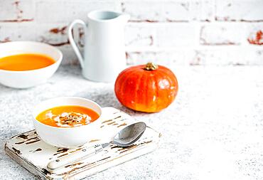 Homemade pumpkin cream soup served in white ceramic bowl on white table with spoon decorated with whole pumpkin, angle view, selective focus. Autumn cozy comfort food, background space for text