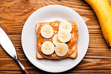 Toast with peanut butter and banana on white ceramic plate with knife on rustic wooden background, top view flat lay