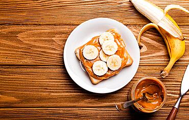 Toast with peanut butter and banana on white ceramic plate with knife on rustic wooden background, top view flat lay, space for text