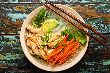 Asian soup with rice noodles, chicken and vegetables in ceramic bowl served with spoon and chopsticks on rustic wooden background from above, Chinese or Thai cuisine