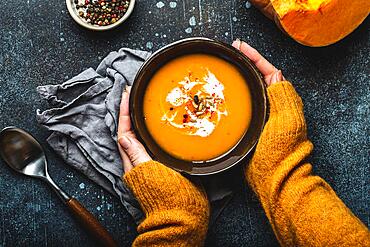 Female hands in yellow knitted sweater holding a bowl with pumpkin cream soup on dark stone background with spoon decorated with cut fresh pumpkin, top view. Autumn cozy dinner concept