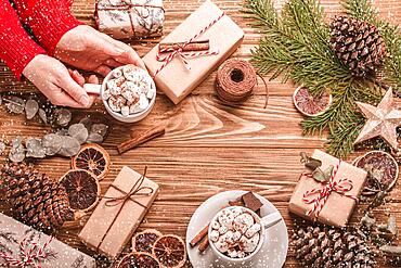 Christmas flat lay with female hands holding a cup of marshmallow cacao, wrapped presents, fir tree branch at wooden background. Xmas decoration top view, cozy festive composition, copy space