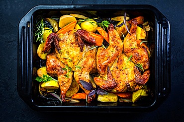 Baked whole chicken with potatoes, carrots and herbs in baking pan on black background