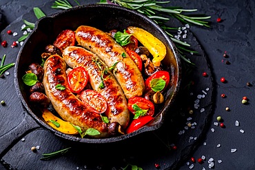 Grilled sausages and vegetables with herbs in iron pan on black background