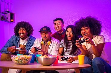 Adult party. Young people sitting on the sofa playing video games with popcorn. Joystick or controller in hand, purple led