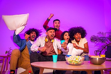 Adult party. Young people sitting on the sofa watching a football game with popcorn. Emotion for soccer, purple led, celebrating a goal