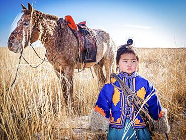 A boy in traditional dress stands next to a horse. Dornod Province, Mongolia, Asia