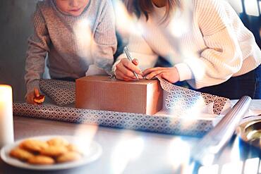 Young family mother and little boy son wrapping packing Christmas gifts for family members while standing at table near beautiful decorated xmas tree at home, kid preparing New Year presents with mom