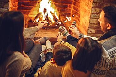 Happy young family parents with two small kids gathering around brick fireplace in cozy and warm country house during Christmas holidays in winter. Father, mother ad children sitting by fire at home