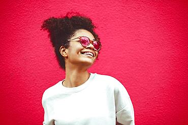 African ethnicity young woman in stylish sunglasses, with curly hair tied up in high ponytail, looking away while smiling broadly showing straight perfect teeth, posing against wall background