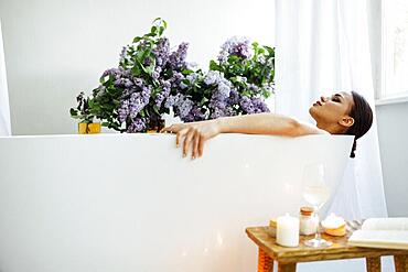 Side view of the young brunette woman reading the book and drinking white wine in bath with foam and candles