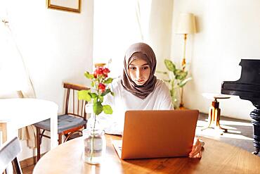 Pretty young middle eastern woman wearing hijab using laptop at home. Muslim girl preparing for entry exams. Home education and online studing concept