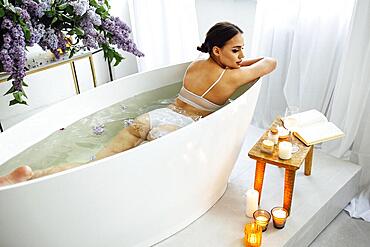 Top view of the young brunette woman reading the book and drinking white wine in bath with foam and candles