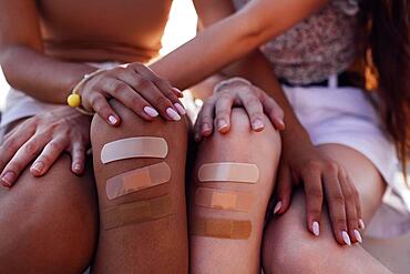 Cropped view of two diffrent races female teen friends with adhesive plasters on injured knee sitting on border bench. Diversity peaple and care concept