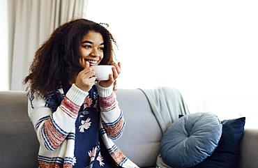 Cheerful smiling african american girl relaxing on couch with cup of coffee, resting at home, enjoying day off, wearing warm clothes