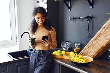 Afro woman holding a vegetables drink. Young african american female drinking green juice with reusable bamboo straw and reading news on mobile phone in loft apartment at the kitchen. Home concept. Healthy lifestyle concept. Copy space