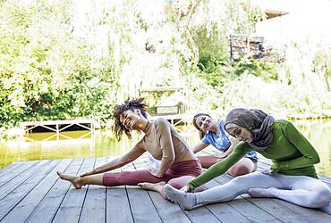 Group of teenages is engaged in fitness on pier by the lake or river. Young and positive girls go in for sports. Muslim in a hijab, african american and white females are doing stretching in the park