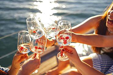 Female hands holding glasses of champagne with raspberries on board the boat. Sunset sky and sea on the background. Making a celebratory toast with sparkling wine. Birthday, holiday, party and friendship concept