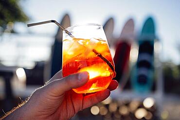 Orange slice and aperol spritz cocktail on a stone against the backdrop of a sandy beach. Man holding slass with iced citrus sparkling mineral water and straw. Copy space