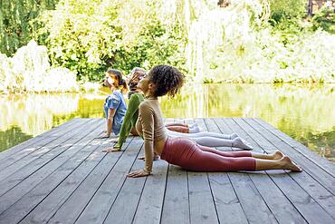 Group of teenages is engaged in fitness on pier by the lake or river. Young and positive girls go in for sports. Muslim in a hijab, african american and white females are doing stretching in the park