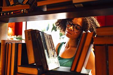 Happy African American female student studing in university library and looking for the book, wearing glasses