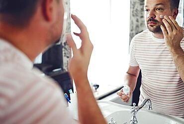 Millennial handsome man looking in mirror, applying moisturizing after shaving cream on cheeks in bathroom, head shot close up. Well groomed young guy doing skincare morning routine after showering