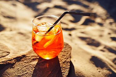 Orange slice and aperol spritz cocktail on a stone against the backdrop of a sandy beach. Glass with iced citrus sparkling mineral water and straw. Long shadows. Top view. Copy space