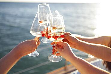 Female hands holding glasses of champagne with raspberries on board the boat. Sunset sky and sea on the background. Making a celebratory toast with sparkling wine. Birthday, holiday, party and friendship concept