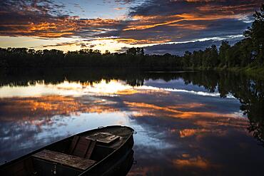 A small boat in a lake at sunset with coloured clouds in orange in the sky. The sky is reflected in the calm lake. Rhine-Neckar District, Baden-Wuerttemberg, Germany, Europe