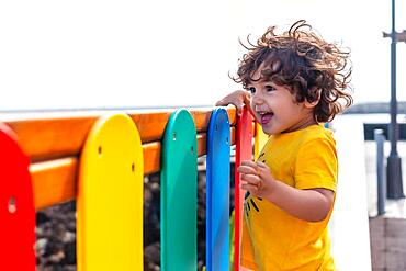 Cute little boy with big eyes and long hair smiling in a game together with the colored barriers of the park